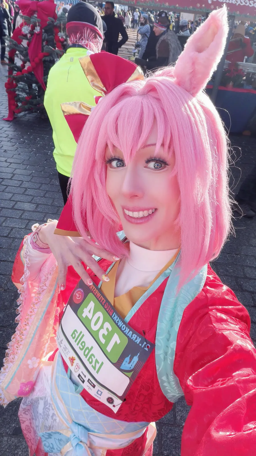 Toraishya in a red and pastel kimono-style outfit with pink hair and horse ears, smiling and showing her race bib on a cobbled square
