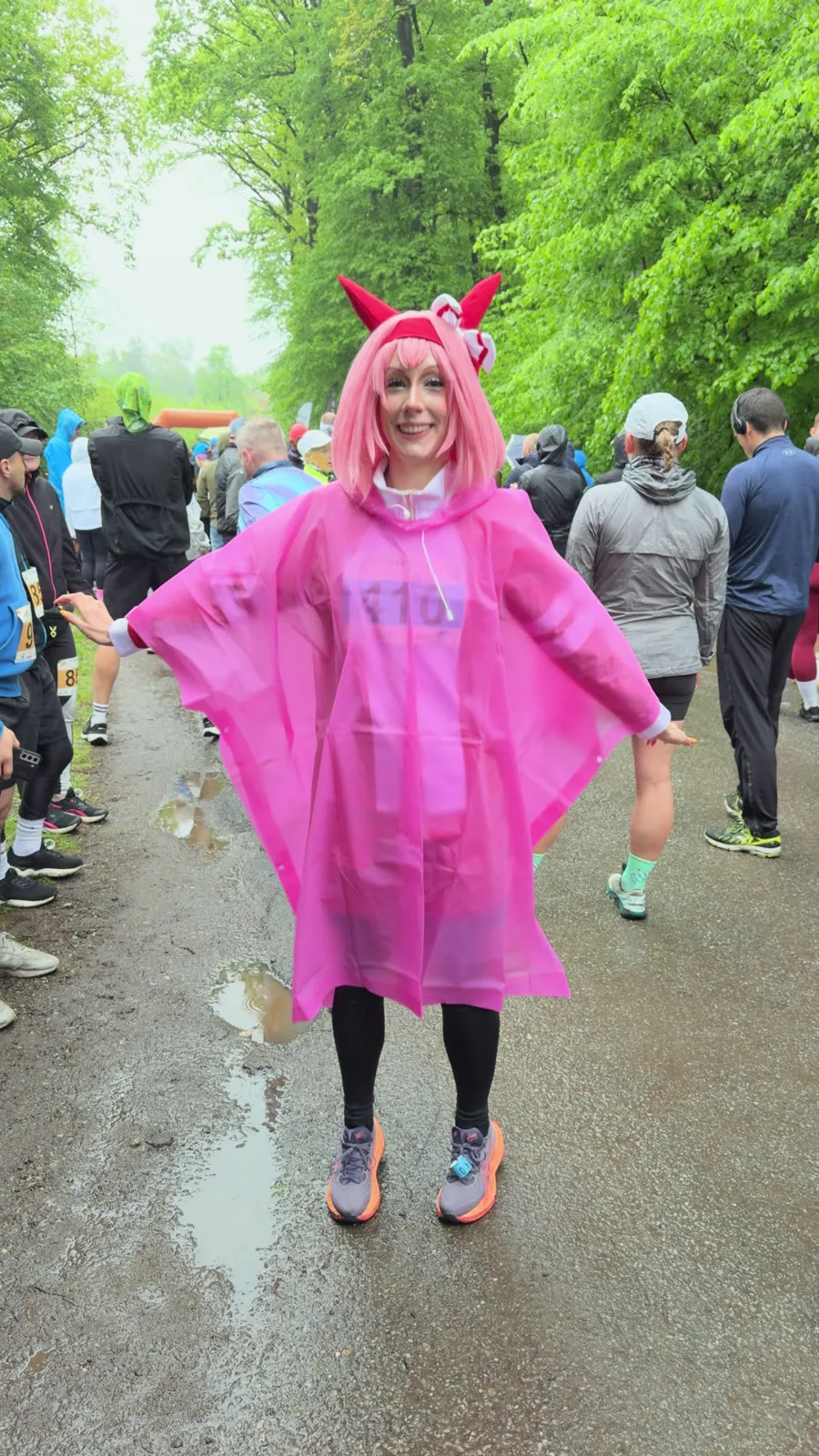 Toraishya in a pink rain poncho and horse ears, arms spread wide on a wet path among other runners